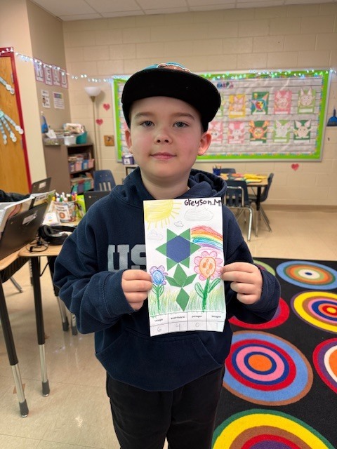 A young student wearing a dark hoodie and baseball cap stands in a classroom, holding up a colorful drawing made of geometric shapes arranged like a flower. Tables, chairs, and classroom supplies are visible behind them.