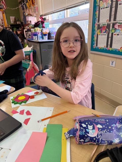 A student with glasses sits at a desk cutting colored paper shapes, with craft materials and a pencil case spread out in front of them.
