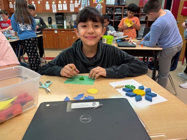 A student smiles at the camera while working with colorful geometric pattern blocks at a desk, with other students working in the background.
