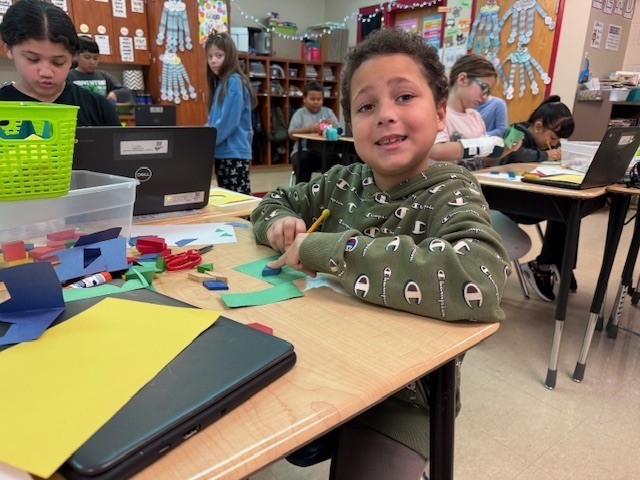 A student sits at a desk drawing or arranging shapes on green paper, with classroom materials and other students visible behind them.