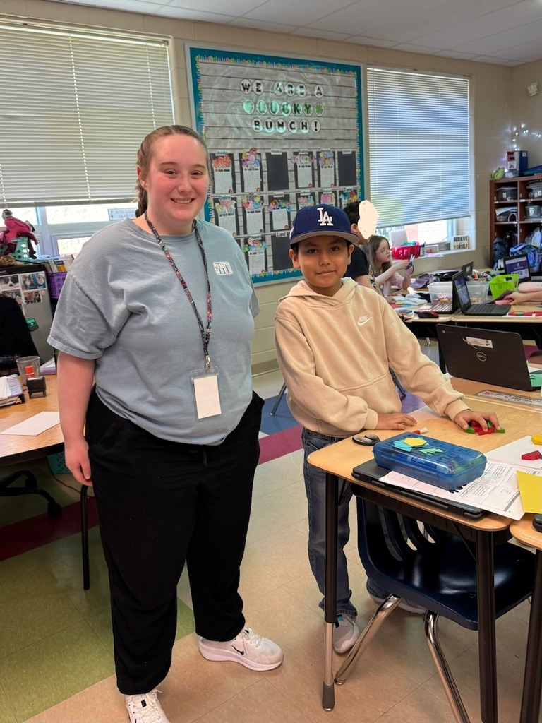 A teacher stands smiling beside a student wearing a blue LA cap at a desk with school supplies and a laptop in a classroom.