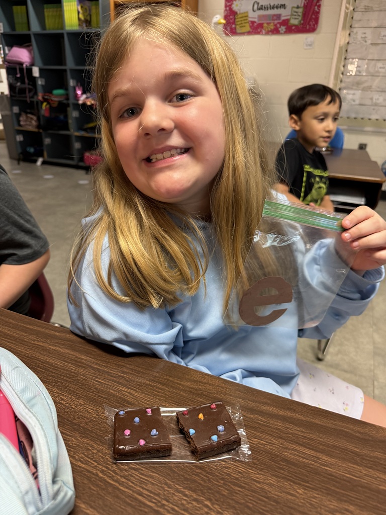 A smiling young girl in a classroom holds up a small plastic bag with a lowercase letter “e” inside. She is seated at a desk with two frosted brownies topped with colorful candy pieces in front of her. Another child and classroom shelves are visible in the background.