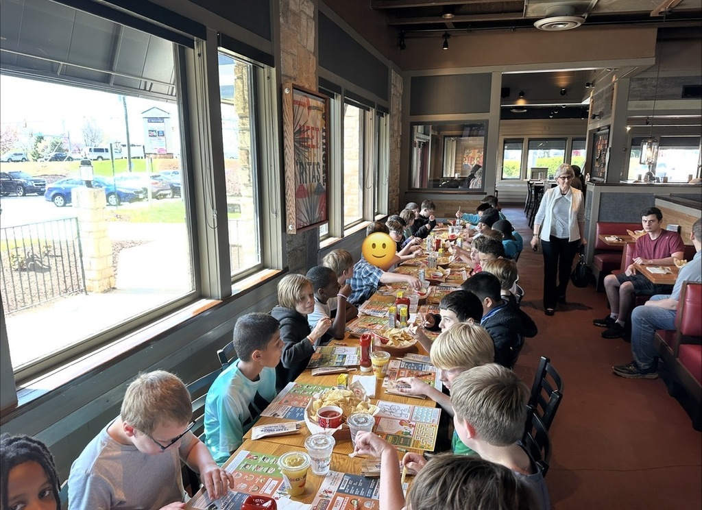 Another view of the long table shows children eating and talking together while an adult walks nearby, with sunlight coming through the windows.