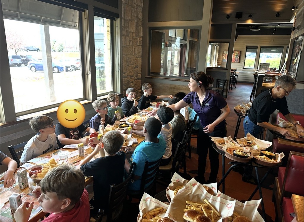 A server delivers multiple trays of food—fries, macaroni and cheese, pizza, and chicken tenders—while adults and children sit in red booth seating nearby.