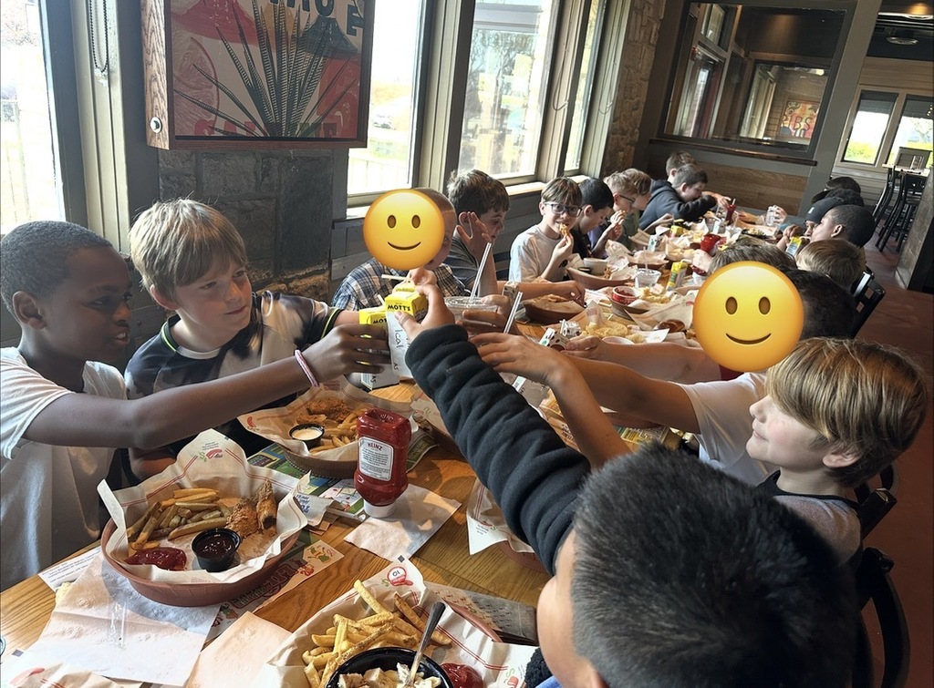 A group of children seated at a long table raise drinks together for a toast while eating fries and chicken tenders.
