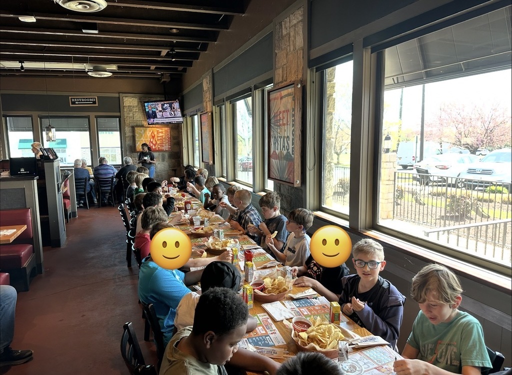 A long table of children eat together in a restaurant, with baskets of chips, drinks, and meals spread out in front of them.