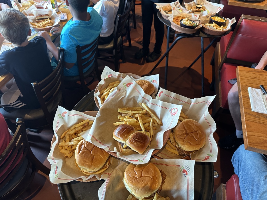 Trays of burgers and fries are arranged on a server stand in a busy restaurant, with children seated at tables in the background.