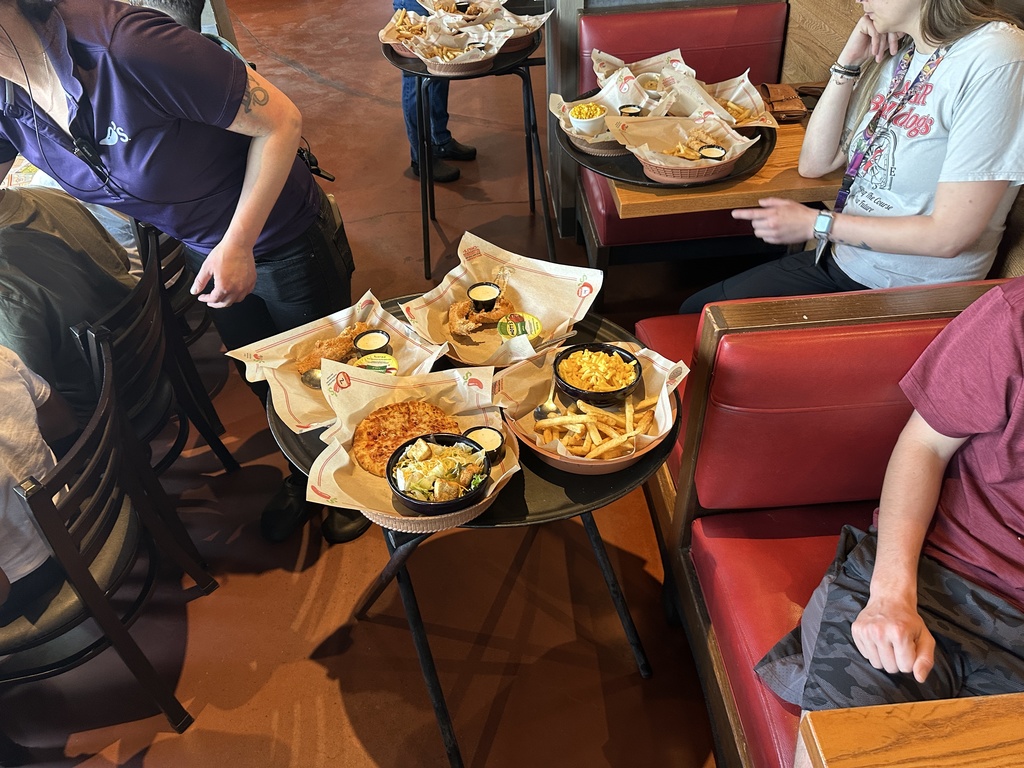 A restaurant staff member hands out food to children seated along a long table by large windows, with trays of meals nearby.