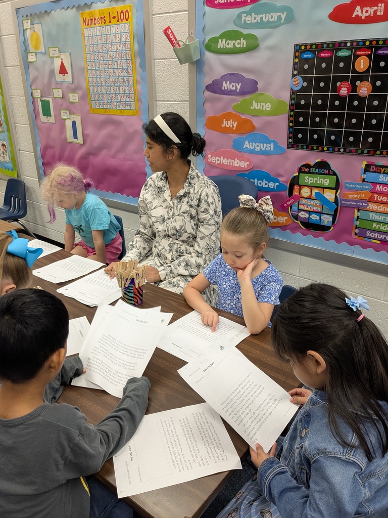 Several elementary students sit around a table reading printed worksheets together. An adult sits with them, guiding the activity. The classroom wall behind them displays a calendar, months of the year, and colorful educational decorations.