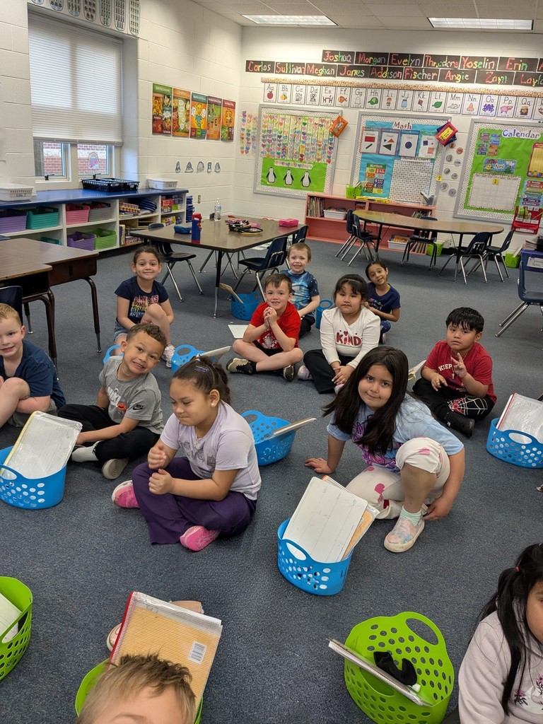 A group of young elementary school students sit on a classroom carpet in a circle, smiling and looking toward the camera. Each child has a small basket and a notebook or whiteboard. The classroom walls are decorated with colorful posters, alphabet letters, and learning charts, and tables and chairs are arranged around the room.