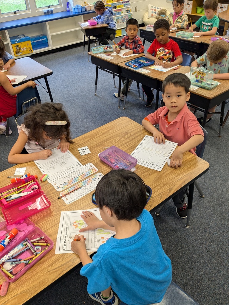 Elementary students sit at desks in a classroom, working independently on worksheets and coloring activities. Crayons, scissors, and pencil boxes are on the desks. The students appear focused, and classroom shelves and materials are visible in the background.