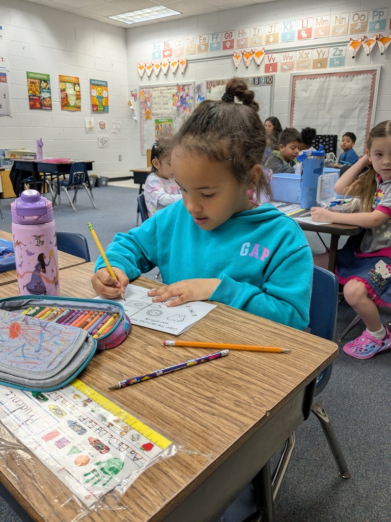 A young student sits at a desk in a classroom, concentrating while drawing or writing on a worksheet with a pencil. Crayons and school supplies are spread across the desk. Other students sit at nearby desks in the background, also working.