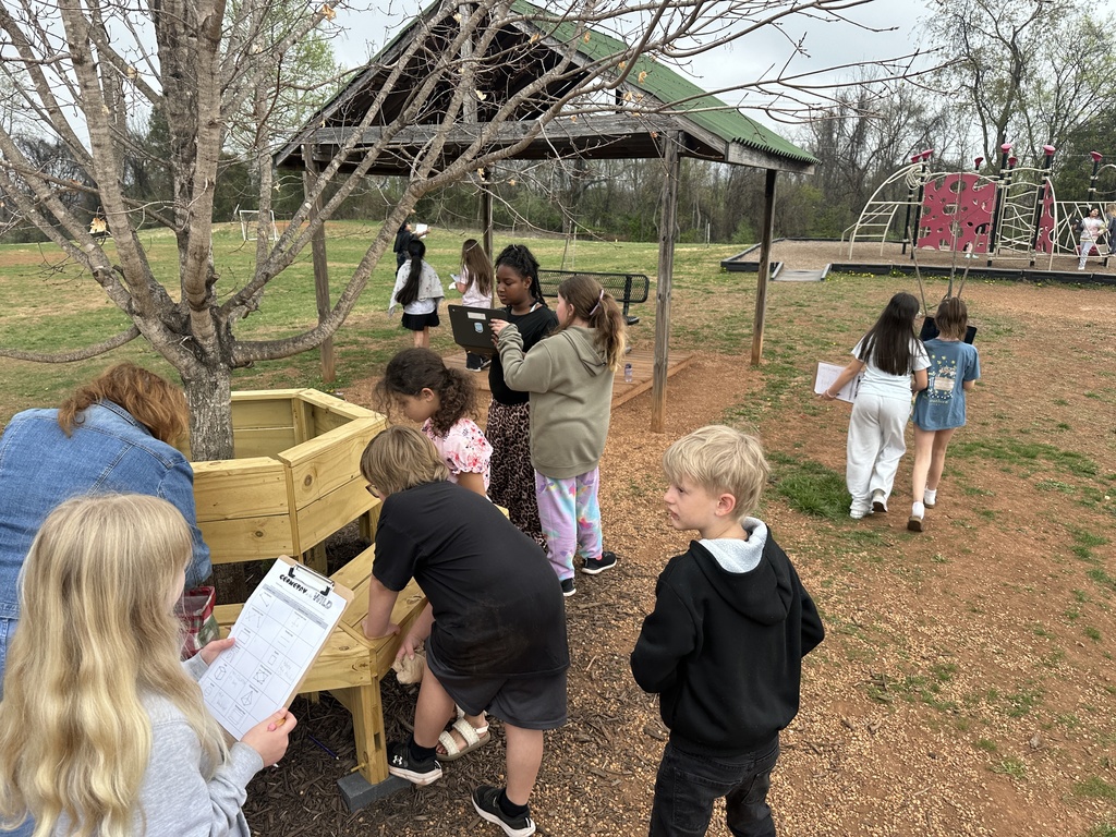 Students work together outdoors near a wooden garden bed and pavilion, using clipboards and a laptop to complete a geometry scavenger hunt on a school playground.