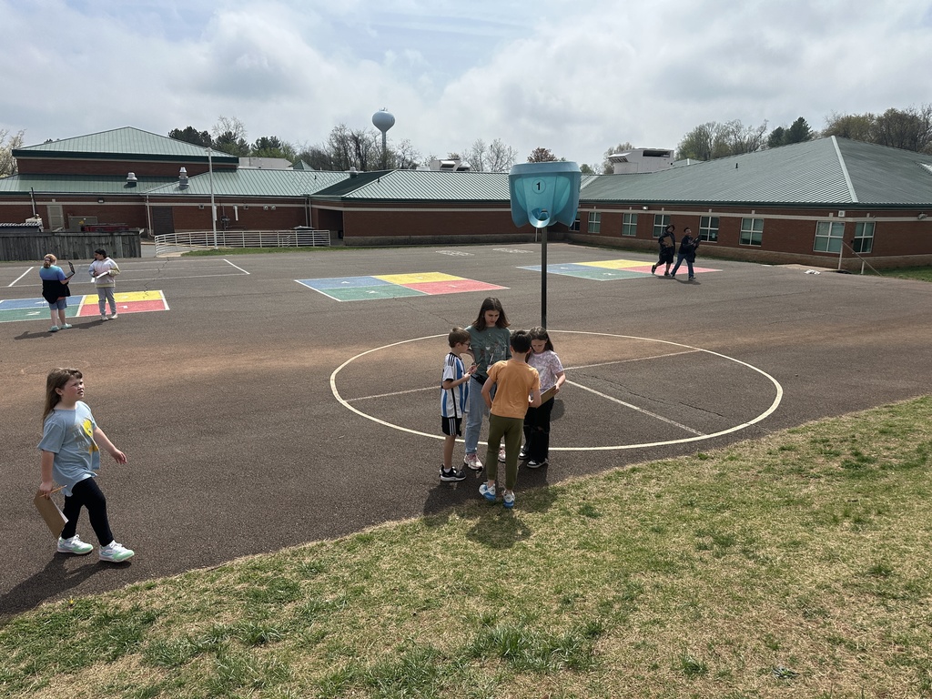 Wide view of students gathered on a playground court with colorful painted squares, collaborating in small groups on a geometry activity.