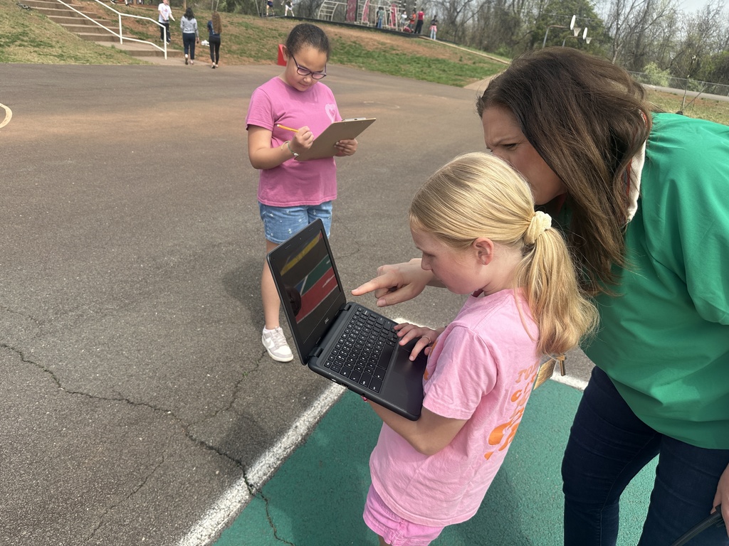A teacher leans in to assist a student using a laptop on the playground, while another student in the background writes on a clipboard during the activity.  If you want, I can also tailor these for accessibility standards (e.g., shorter, screen-reader optimized versions or add grade-level context).