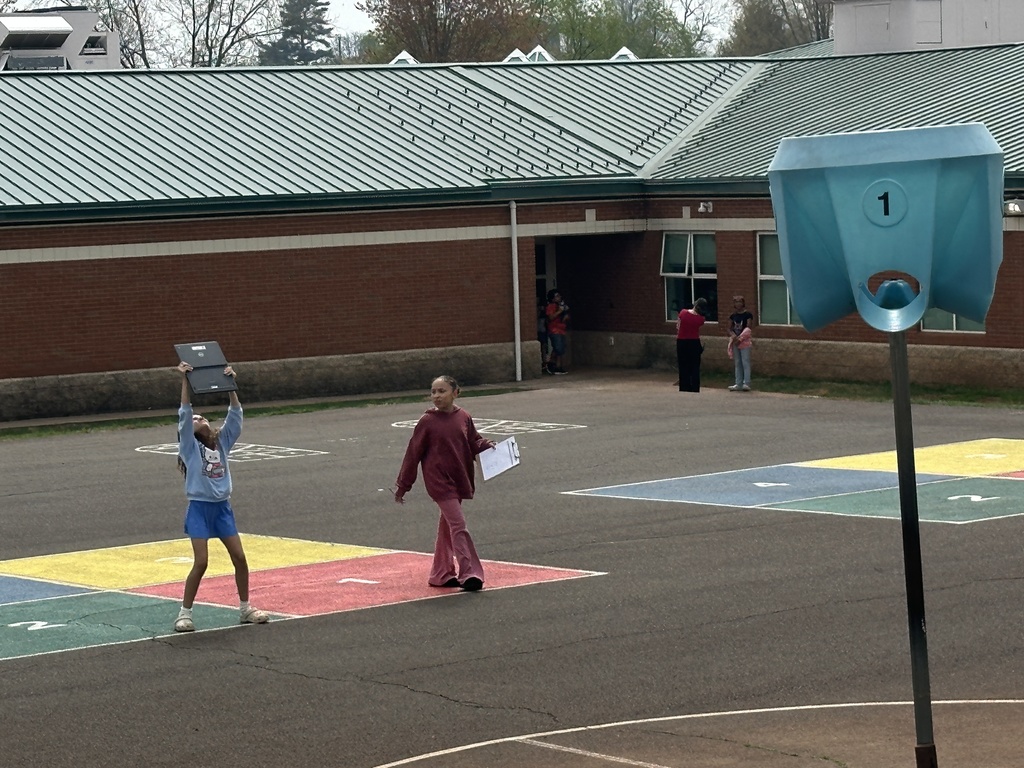 Students explore a blacktop playground with painted shapes while holding clipboards and a laptop, searching for geometric figures around the school building.