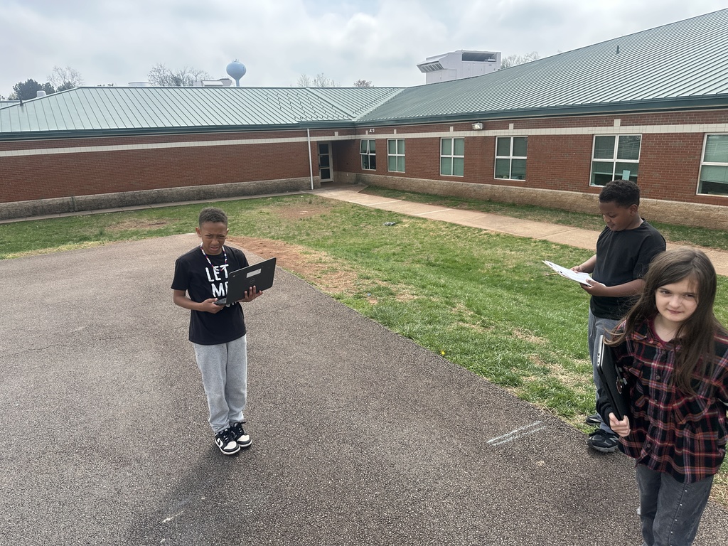 Three students stand on a paved area near the school, each holding a clipboard or laptop, observing and recording geometric shapes in their surroundings.