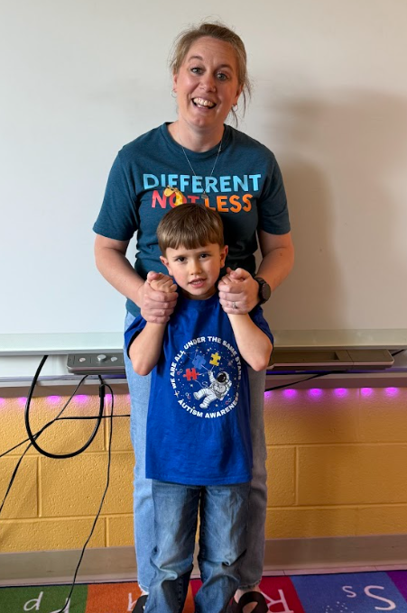 “Teacher and young student wearing blue stand together in a classroom, smiling. The teacher’s shirt reads ‘Different Not Less’ in support of Autism Awareness.”