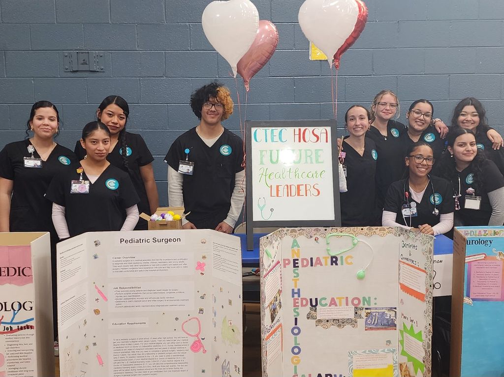 CTEC HOSA students pose behind a display table at Sycamore Park STEM Night, showcasing projects on healthcare careers such as pediatric surgery, anesthesia, and neurology.