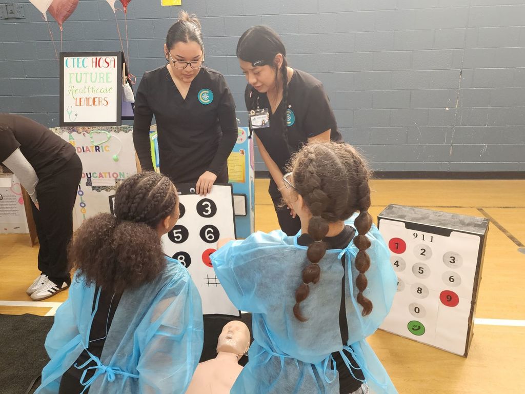 CTEC HOSA students demonstrate a hands-on activity at Sycamore Park STEM Night, guiding two younger students using a large model keypad and a CPR training mannequin while teaching basic emergency response skills.