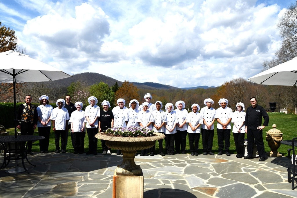 Group of CTEC culinary students and chefs standing outdoors at The Inn at Little Washington, wearing white chef coats and hats, posed on a stone patio with mountains and spring foliage in the background during a class visit.