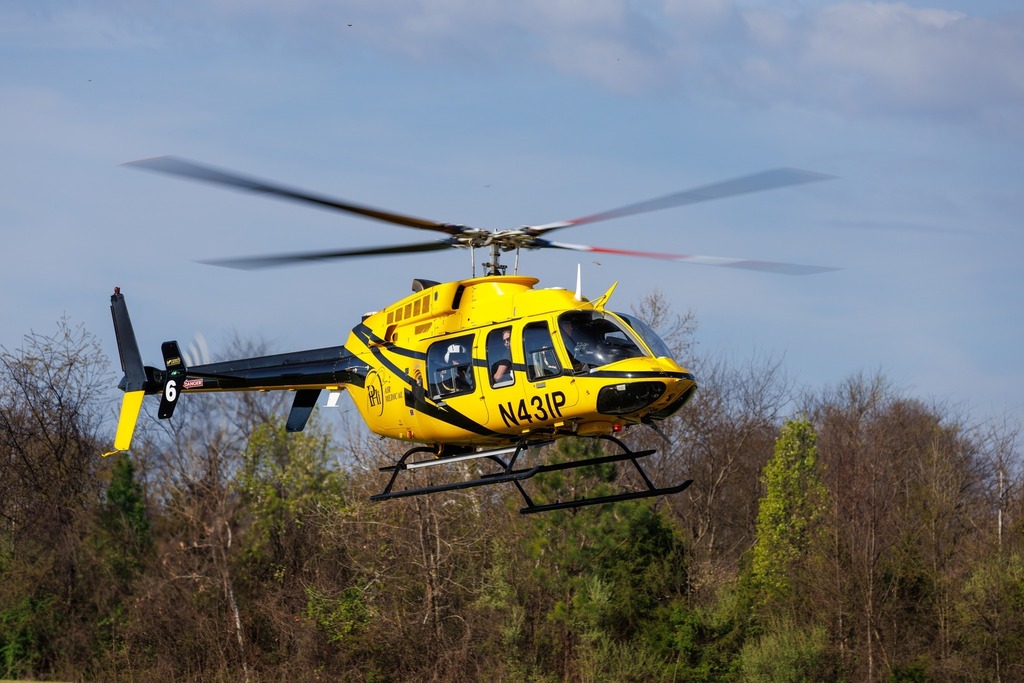 AirCare 6 medical helicopter hovers just above a grassy area, with its rotors spinning and crew visible inside the bright yellow aircraft.
