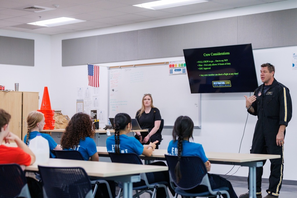 A flight crew member presents to EMT students in a classroom, discussing crew considerations while a slide is displayed on a screen at the front of the room.