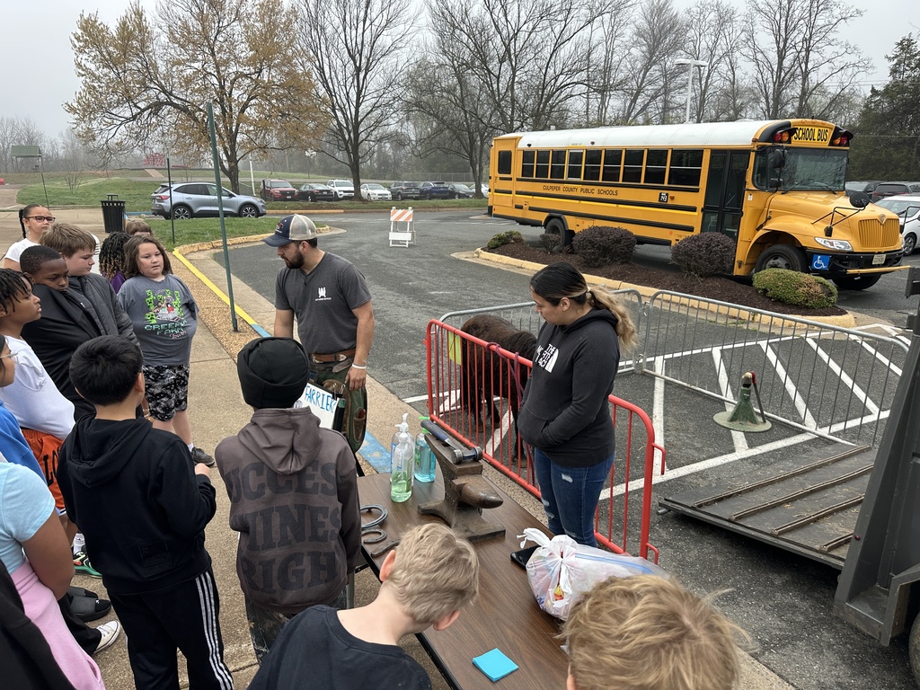 Students gather outside near a school bus as two adults demonstrate tools on a table, related to farrier work.  The setting appears to be a school parking lot.