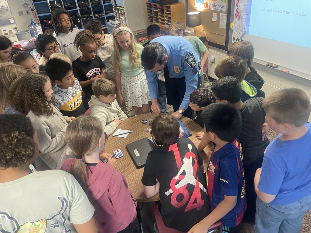 A police officer leans over a table in a classroom, demonstrating an activity to a group of students who are gathered closely and watching with interest.
