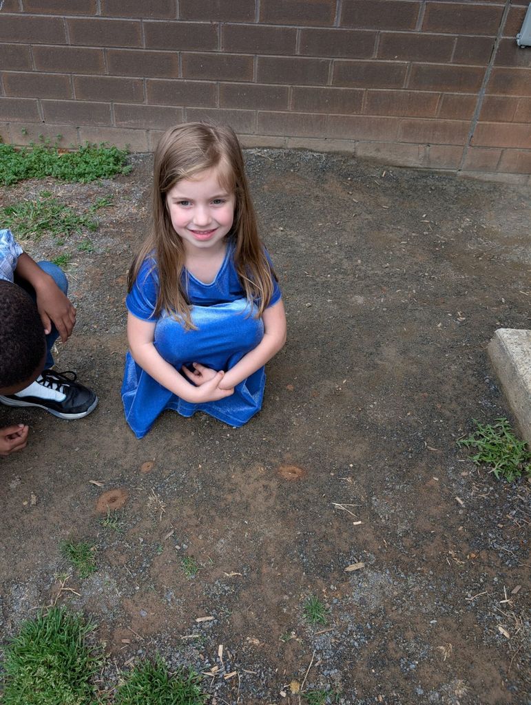 A young student in a blue dress kneels on the ground outside near a brick wall, smiling at the camera while other children sit nearby.