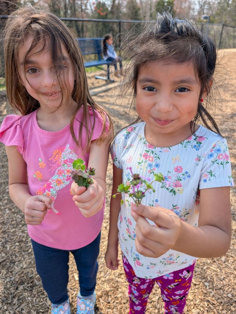Two students stand outdoors smiling and holding small flowers or plants up toward the camera.