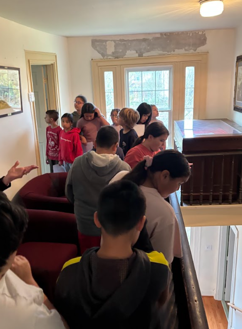 A group of students stands closely together inside a historic building, gathered near a staircase and windows as they listen and look around.