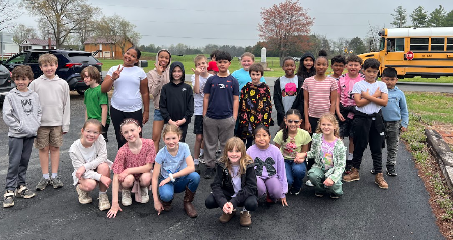 A large group of students pose outdoors on a paved area, smiling at the camera with a school bus and grassy field in the background.