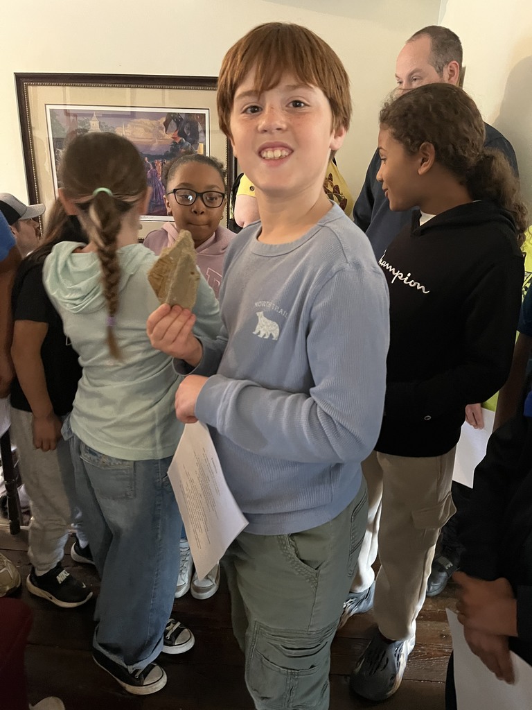 A student smiles at the camera while holding a small artifact, surrounded by classmates inside a historic room.