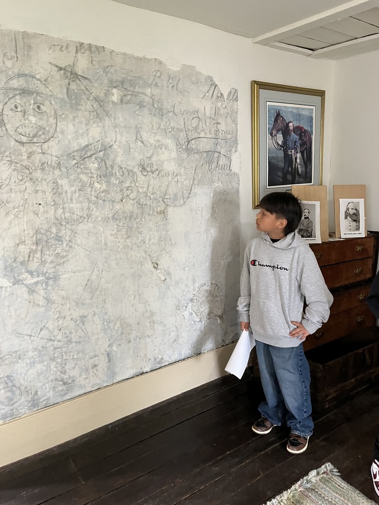 A student stands indoors looking at an old wall covered with faint drawings and writing, holding a paper in one hand.