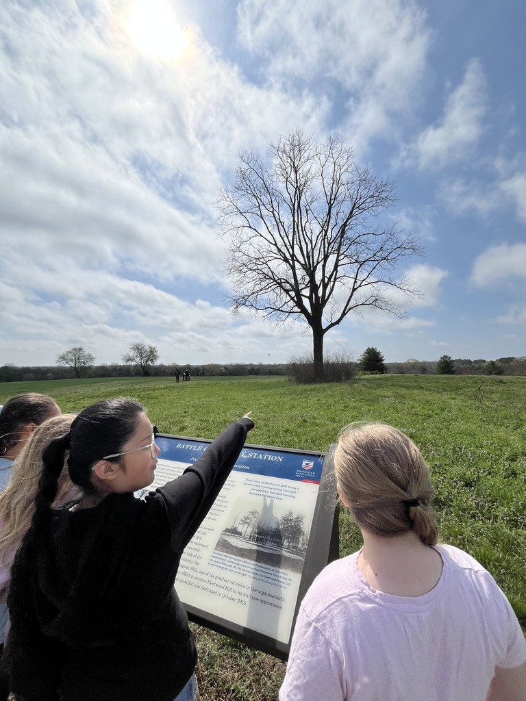 Students stand at an outdoor historical site, reading a sign and pointing toward a large tree in an open field under a partly cloudy sky.