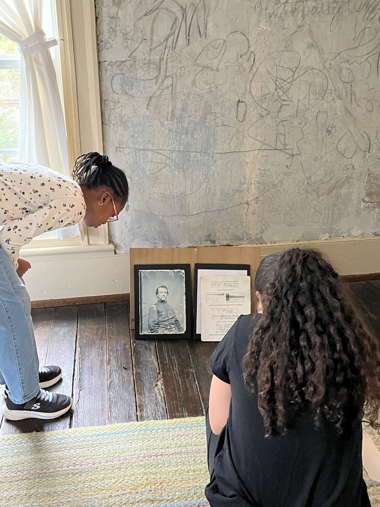 Two students kneel on a wooden floor, closely examining framed historical photos and documents leaning against a wall with old markings.