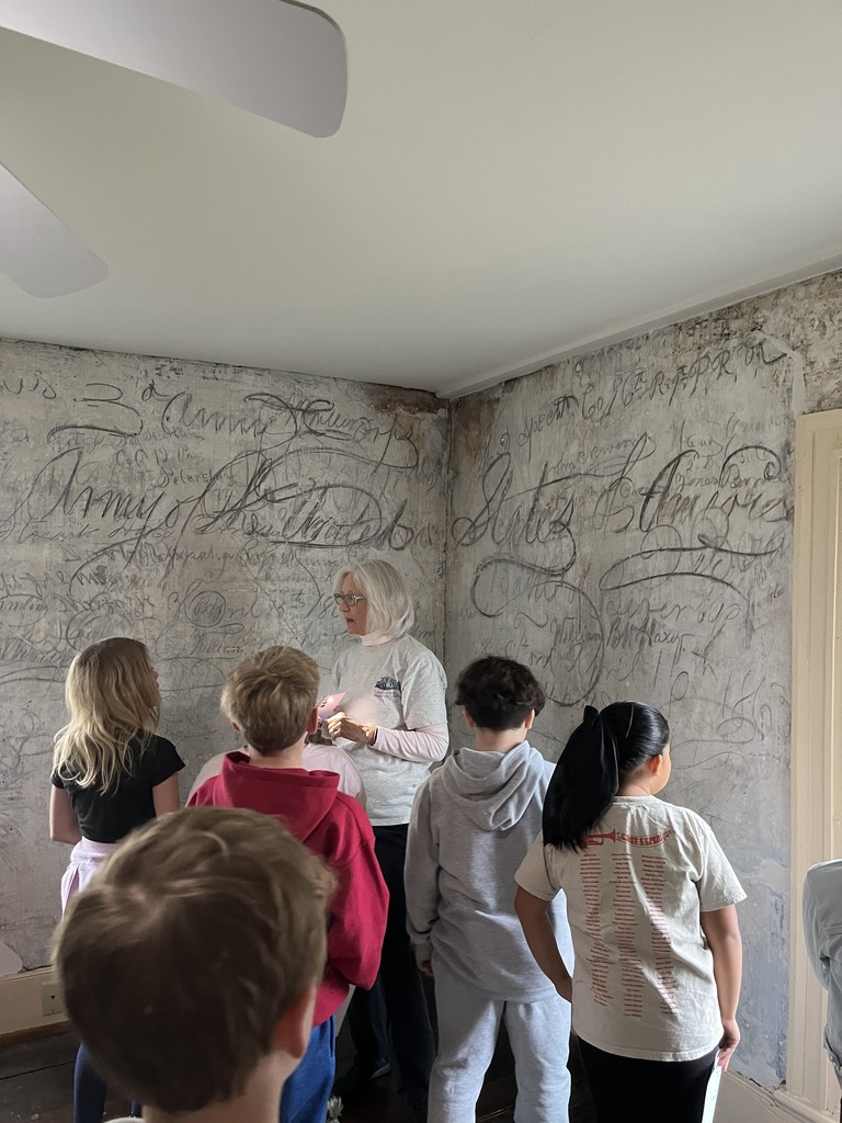 A group of students listens to an adult guide inside a room with walls covered in historic signatures and writing.