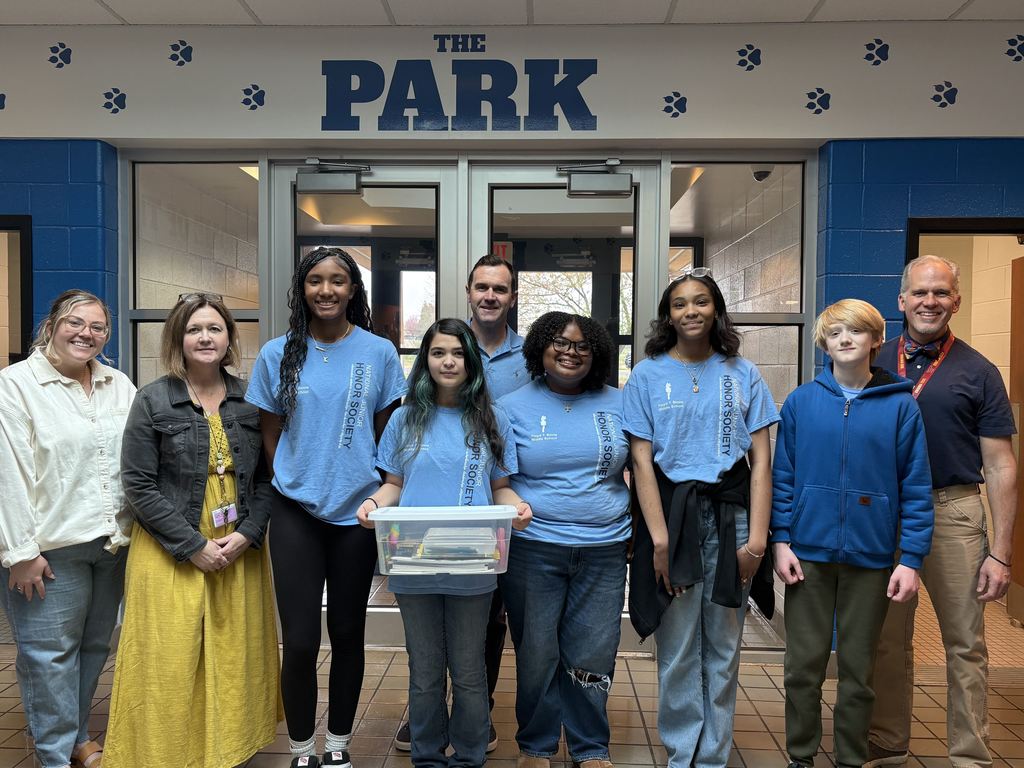 A group of eight people—four adults and four students—stand together inside a school entrance under a sign that reads “THE PARK,” with blue paw prints decorating the wall. The students in the center wear matching light blue “Honor Society” shirts, and one student holds a clear plastic bin filled with books or supplies. The adults on either side are dressed in casual-professional clothing, and everyone is smiling at the camera. The setting appears to be a school hallway or entryway with glass doors behind them.