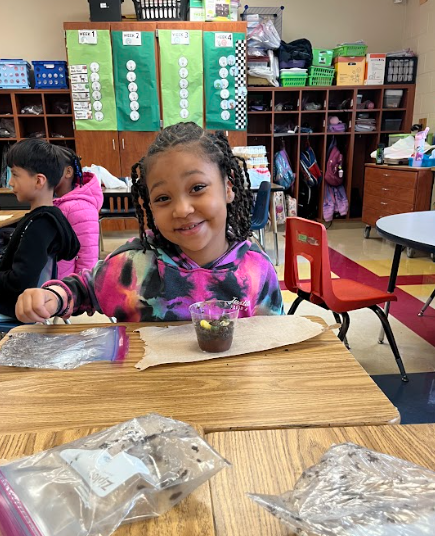 A smiling student sits at a desk with a small cup of dirt and gummy worms, showing their completed project while classmates work in the background.
