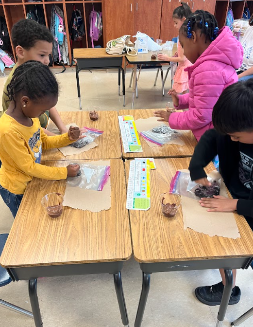 Several students sit at grouped desks, working together to mix soil in bags and create dirt cup projects.