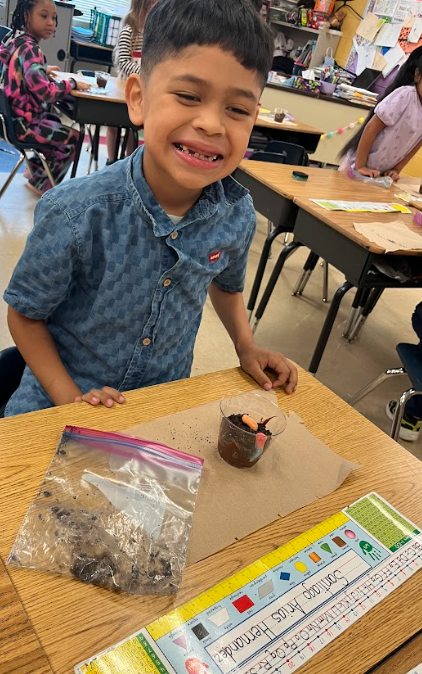 A student smiles at the camera while sitting at a desk with a cup of dirt and gummy worms, alongside a plastic bag of soil and classroom materials.