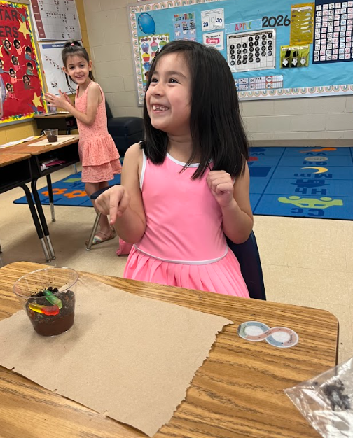 A smiling student in a pink dress sits at a classroom desk, pointing excitedly at a small cup filled with dirt and gummy worms, while another student stands behind her.
