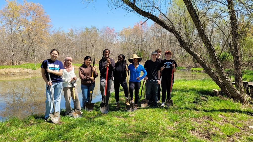 A group of students stand side by side on the grassy bank of a calm stream, smiling and posing with their shovels after their work. They are dressed casually in T-shirts, hoodies, and jeans, and appear proud and relaxed. Behind them, the water reflects the trees and blue sky, highlighting a bright, sunny day and a sense of accomplishment after planting.