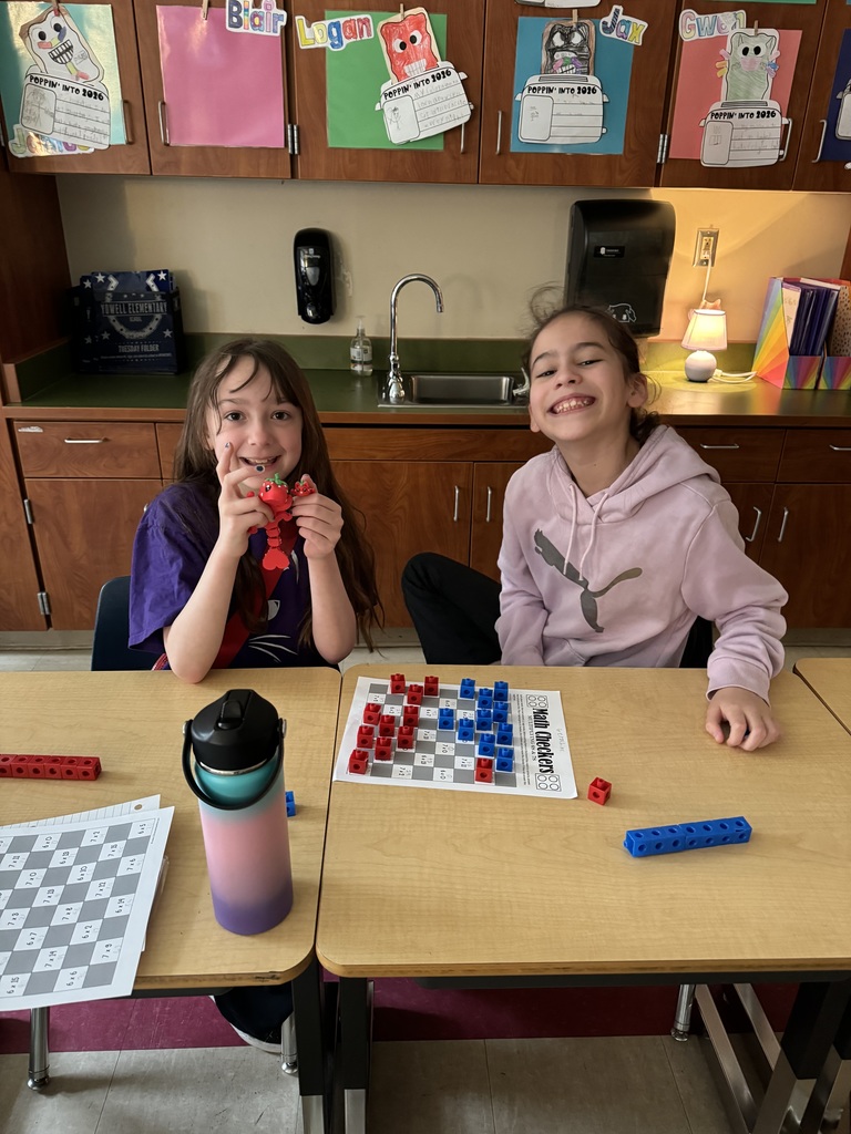 Two girls sit at desks smiling, playing a math-themed board game with red and blue cubes.