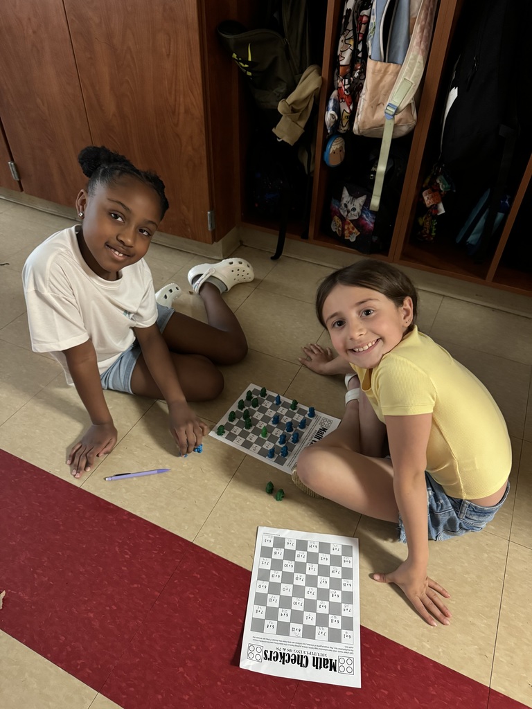 Two girls sit on the floor playing a math board game with small pieces, smiling up at the camera.