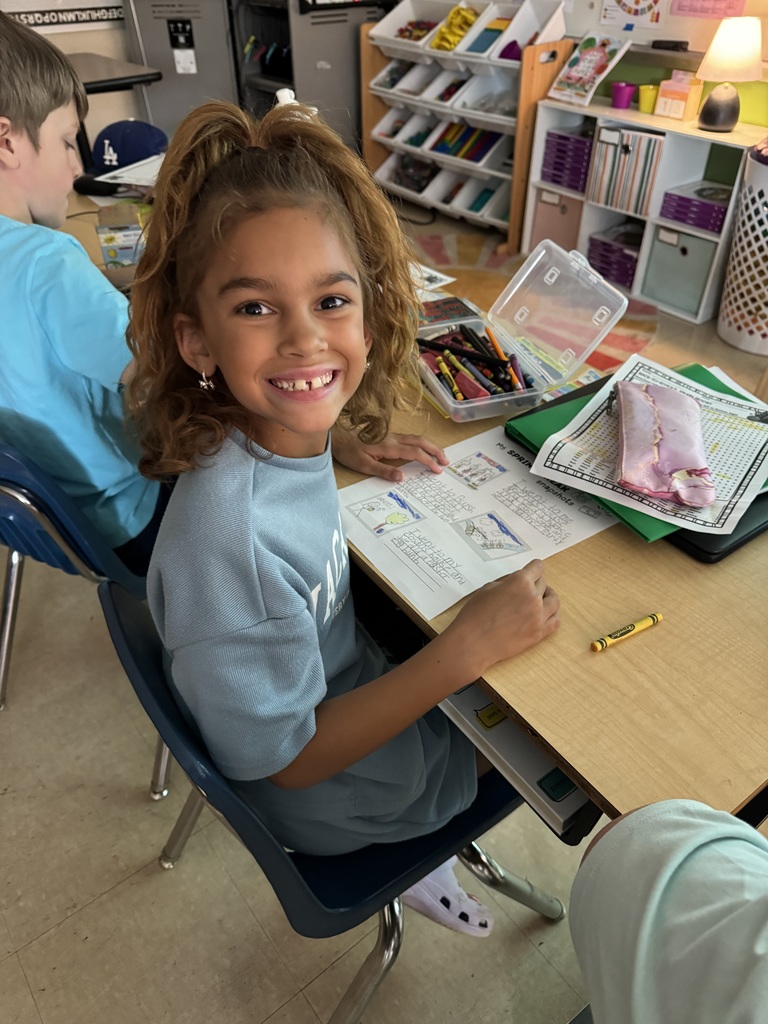 A smiling young girl with curly hair sits at a desk in a classroom, holding a worksheet and surrounded by crayons and school supplies.