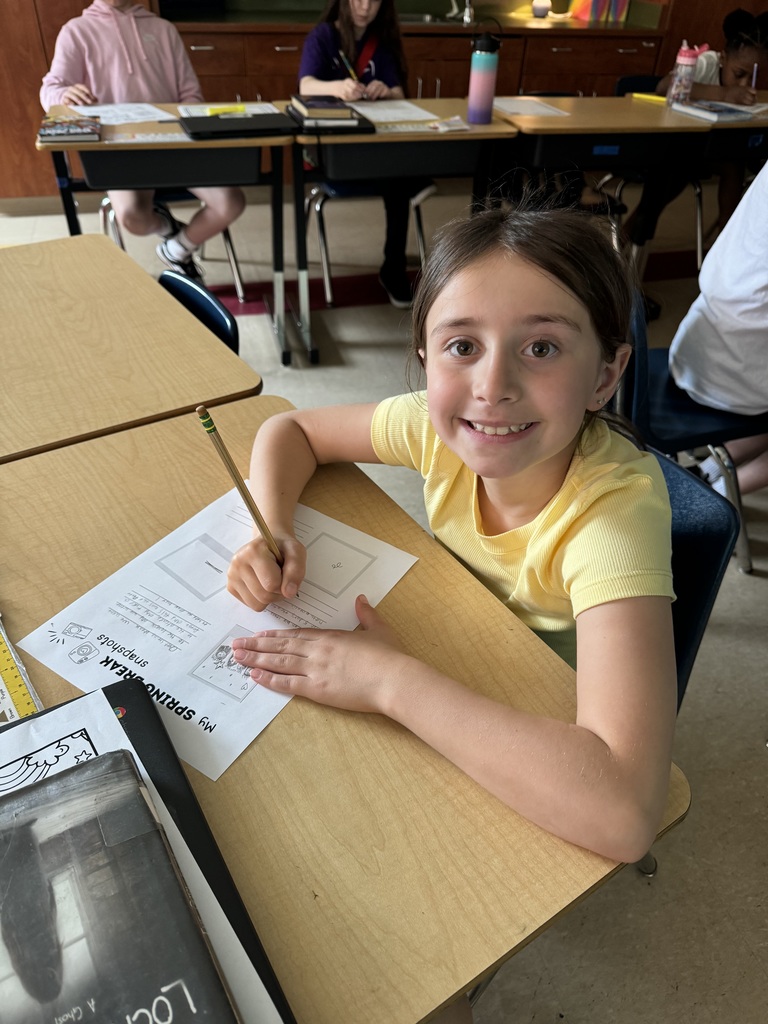 A girl in a yellow shirt smiles while writing on a worksheet at her desk in a classroom, with other students working behind her.