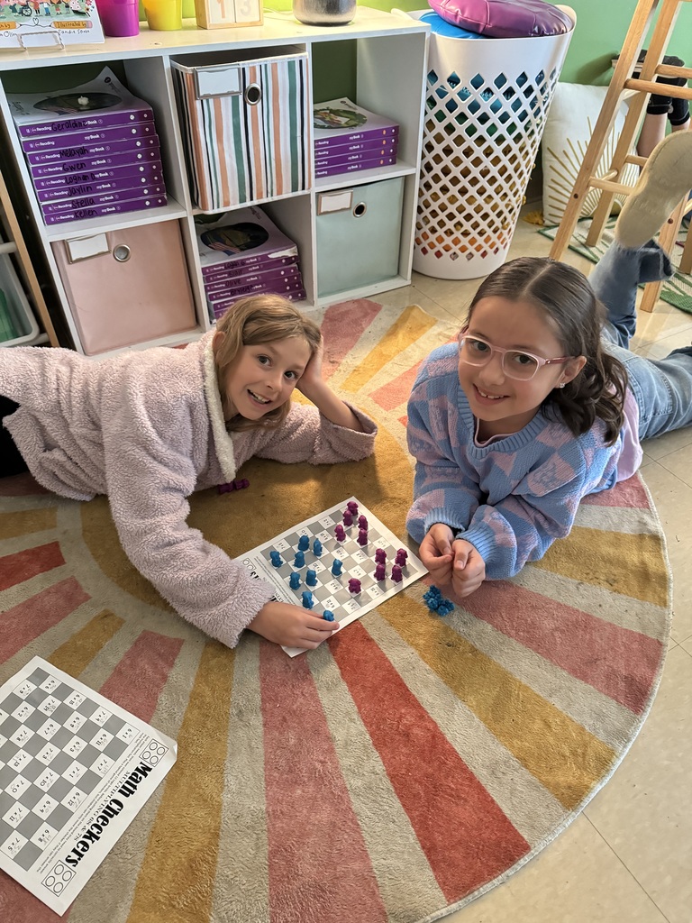 Two girls lie on a colorful rug, smiling while playing a board game with purple and blue pieces.