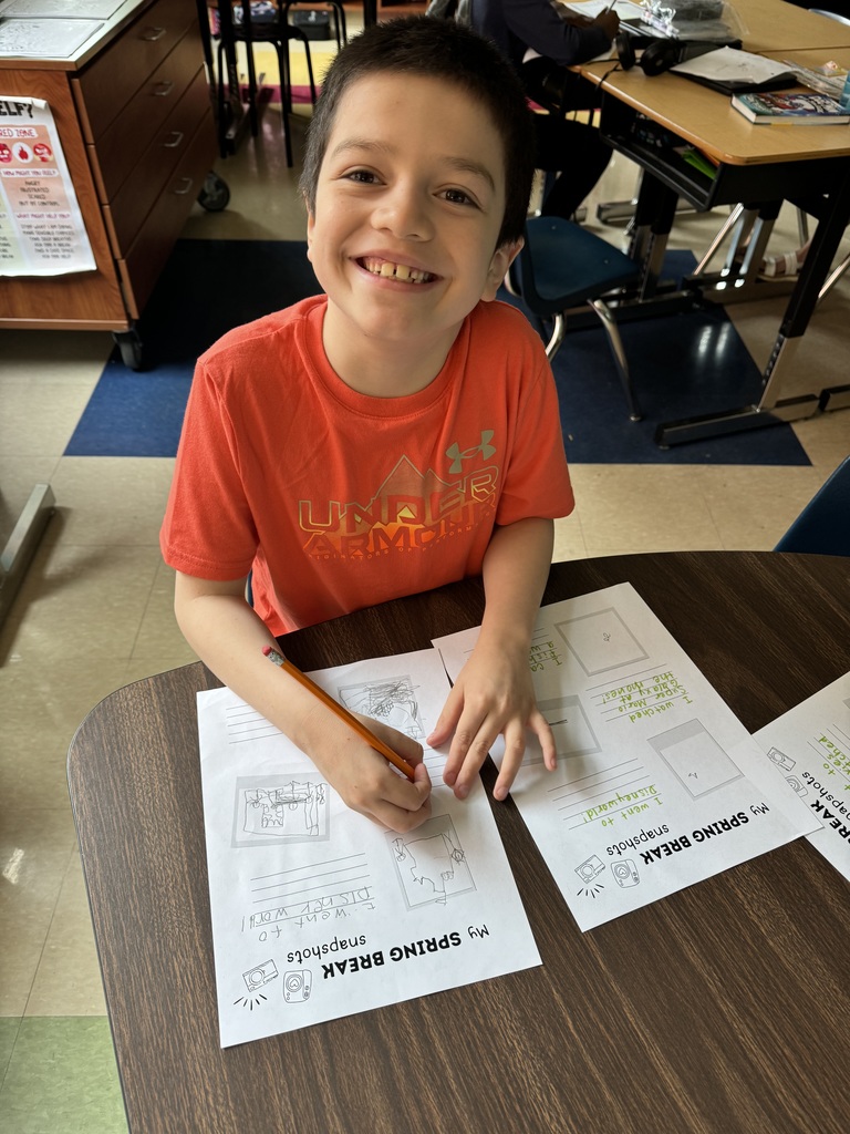 A boy in a bright orange shirt smiles while drawing on a worksheet at a classroom table.